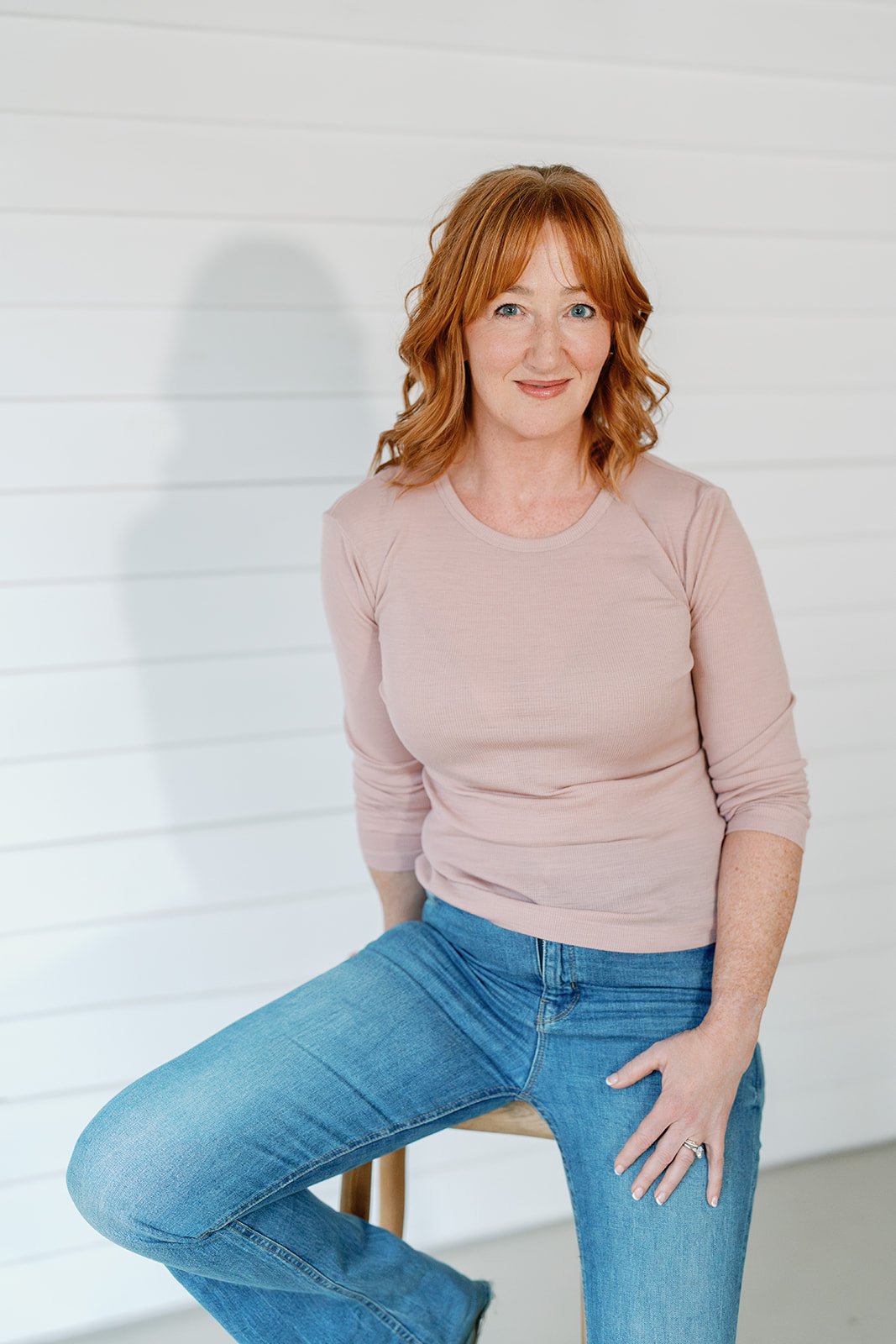 Amanda Alexander smiling, sat on a stool in a blush pink jumper and jeans